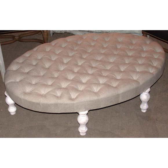 A large oval tufted poof upholstered in a beige linen fabric standing on white patina wooden legs, newly constructed from old and new parts.
