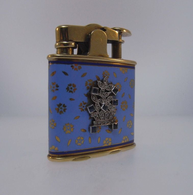 A charming enamelled 14kt gold lighter by Ronson, with a decorative motif of five-petalled flowers on a blue ground.  A central plaque of 7 sapphires and 26 diamonds set in platinum depicts a basket of flowers.  In full working condition.