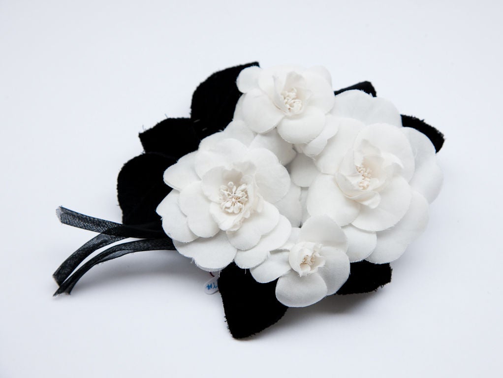 Chanel signature white Camillia pin. This piece is made up of 4 Camillia flowers clustered together making it a rather impressive scale. Flowers are surrounded with black velvet leaves and silk organza stems. Tiny gold Chanel charm authenticates the