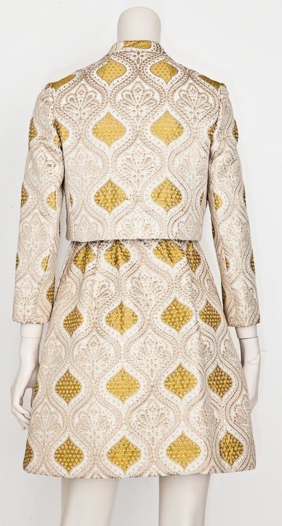 Ivory and gold lame brocade sleeveless dress with matching cropped jacket, designed by Kiki Hart. Dress has a fitted bodice and slightly gathered  skirt. Fabric is an Indian inspired print brocade with an ivory ground and a chartreuse paisley like