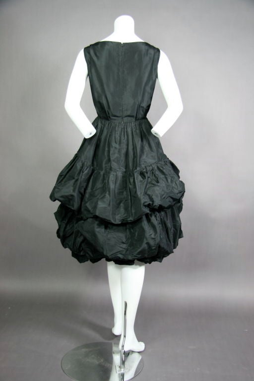 Black Silk Tafetta gown Autumn/Winter 1959. Scooped boat neck, sleeveless. Fitted bodice with pleats to waist. Drop waist and 3x tiered bubble skirt.  Hem is shorter in the front and the drop is longer at the back. Double zip, lined with additional