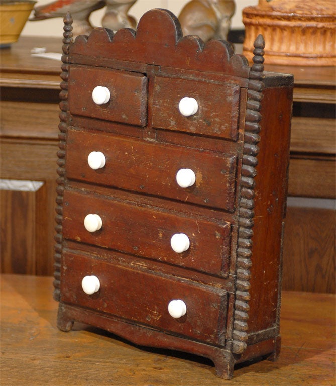 A painted English miniature nest of drawers from the late 19th-early 20th century. This exquisite English miniature brown colored nest of drawers features five drawers with two cubby drawers over three larger ones. The crest is decorated with a