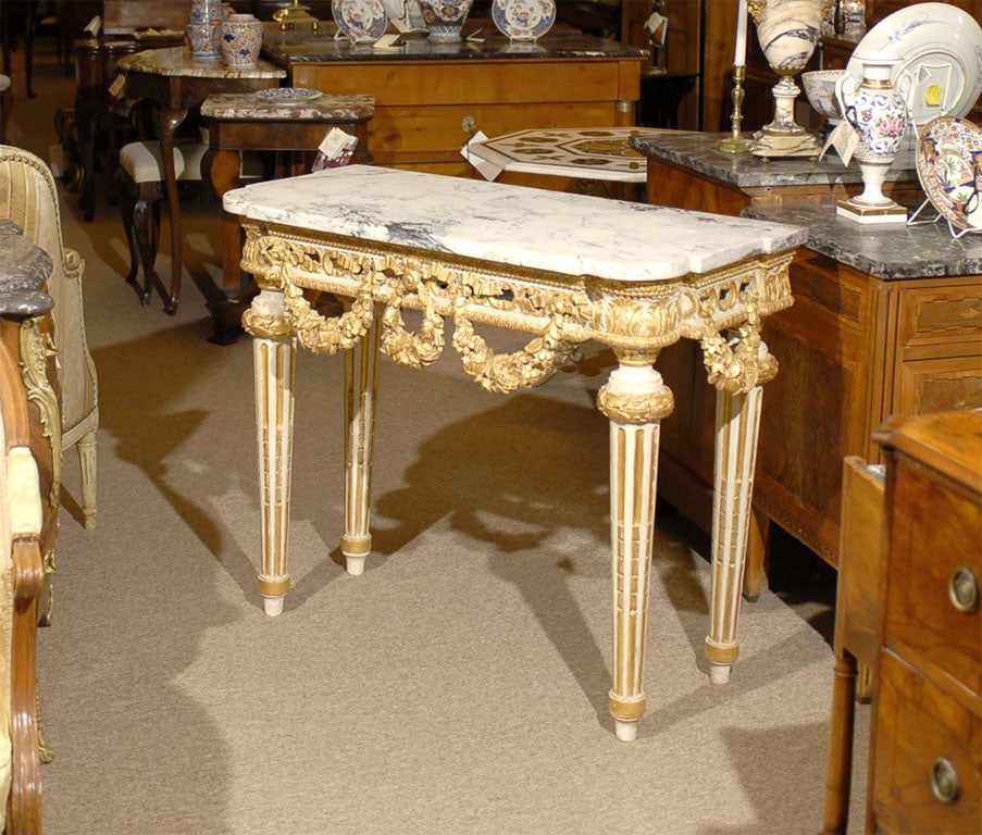 A Louis XVI period painted and parcel-gilt console table in the neoclassical taste, mounted with a marble top. The break-front outline adorned with ribbon and rose garland motifs, and mounted atop four dramatic tapered legs with gilt accents and