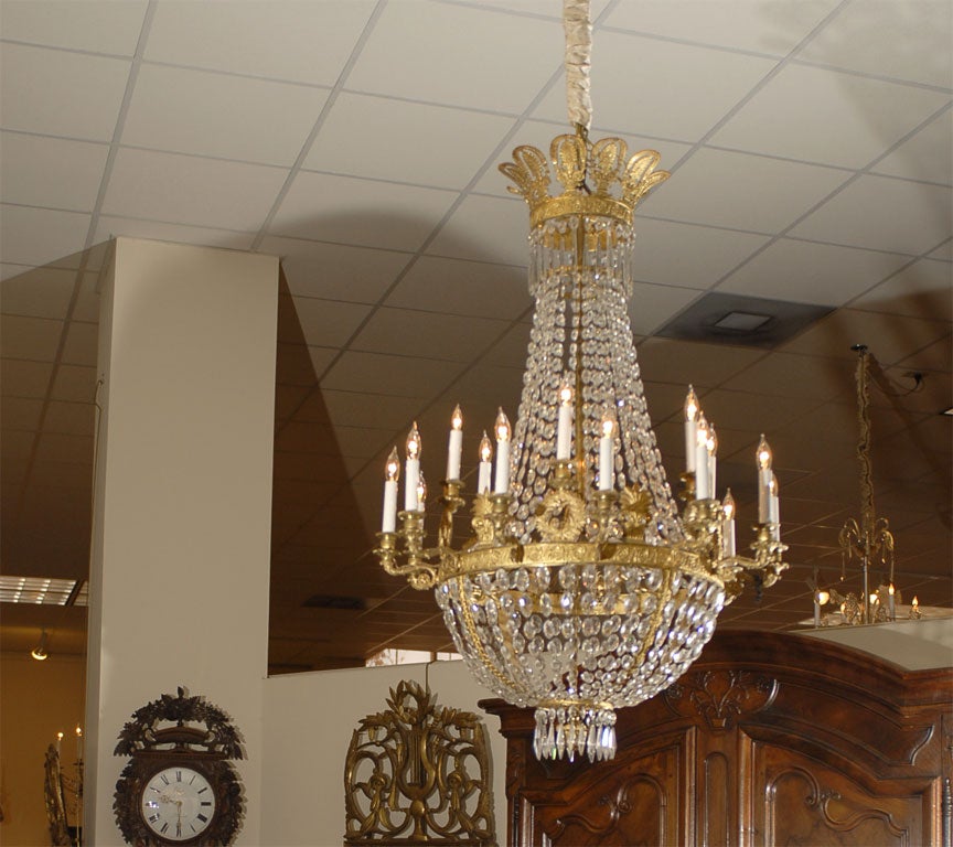 An 18-light chandelier in Gilt-bronze, with strands of cut-crystal beads draping outward from the upper gilt-bronze capital, meeting a two-tiered band of candle-arms. 6 lights on upper tier, each staggered between a pair on the lower tier of arms.