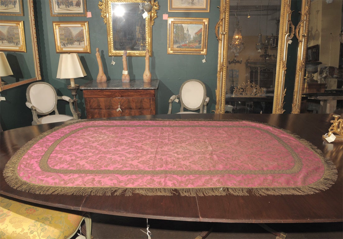 AN OVAL PINK SILK TABLE COVER OVERLAID WITH A FOLIATE PATTERN OF GOLD THREAD TRIMMED IN GOLD METALLIC FRINGE  WITH COTTON BACKING.