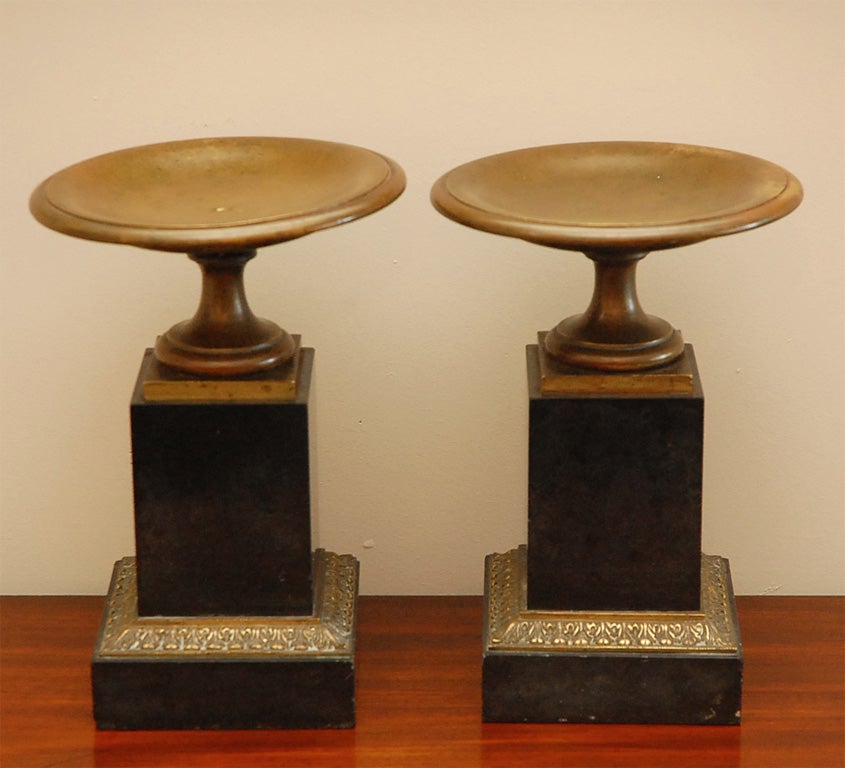 A Pair of Black Marble Tazzas. The round metal tazza tops with incurved stem bases are raised on marble plinths with foliate motif gilt metal on square marble bases.