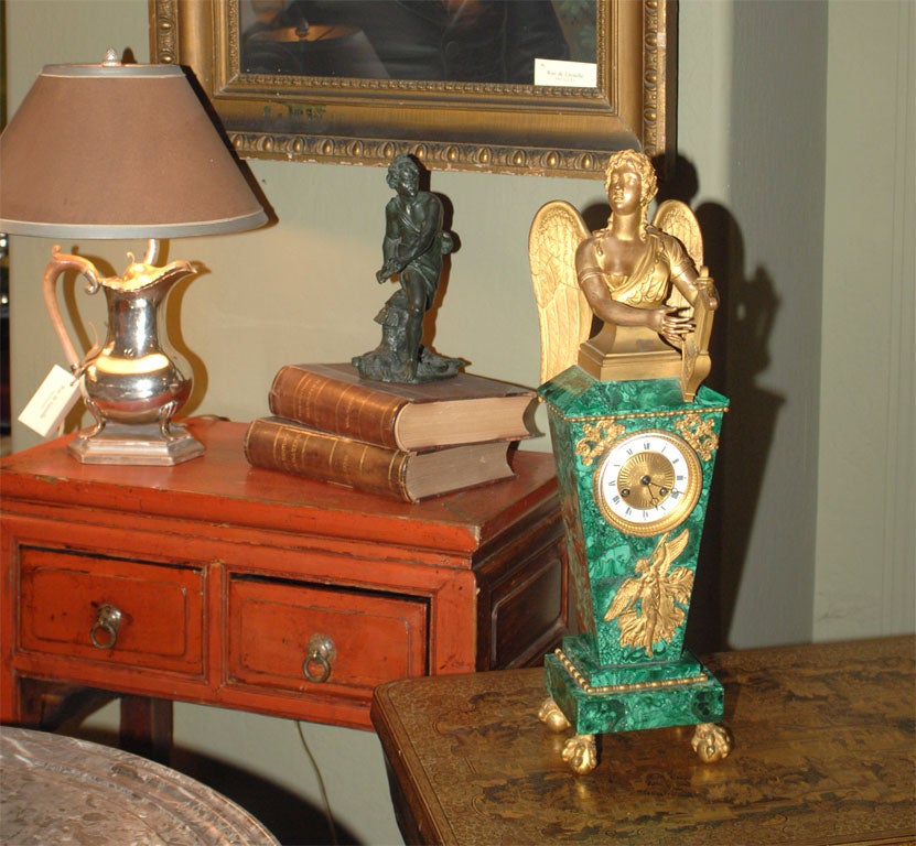 With a gilt bronze muse of music playing a harp atop a base of veneered malachite with ormolu mounts of foliate scrolls and an angel blowing a horn, resting on ball and claw feet.  The base is inset with a clock that is signed, 