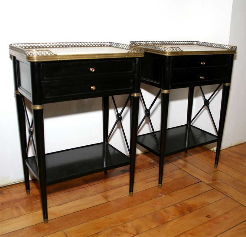 A very chic pair of directoire style end tables with inset white marble tops and gilt bronze openwork galleries.  Each has two small drawers and a lower open shelf.  The legs are round and tapered.