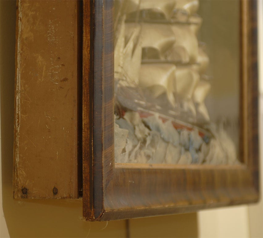 This late 19th century nautical diorama features a three-masted sailboat model under full sail on a stormy sea simulated with painted paper, enclosed in a large grain-painted wooden shadowbox frame. The carved and painted black and red-hulled ship