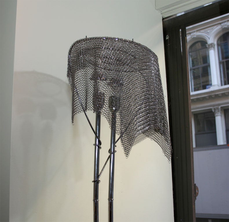 A very sleek and modern floor lamp with two bulbs. The base supports two arms that shoot up connecting in a spiral at the top. The shade of this unique light is a piece of chain maille.