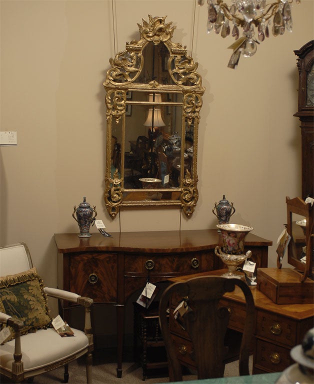 A Regence design gilt-wood mirror, dating from the third quarter of the 1700s, and French in origin. <br />
<br />
The mirror with gilt-wood frame in the early Rococo design, notable for the Trophic crest surmounting the upper section, featuring a