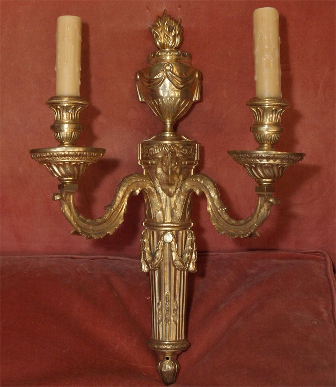Single bronze d'ore Louis XVI applique, having a tapered fluted shaft with a decorative central ram's head and surmounted by an urn with a stylized flame motif.  The shaft supports two scroll arms, terminating in bobeches with nozzles holding waxed