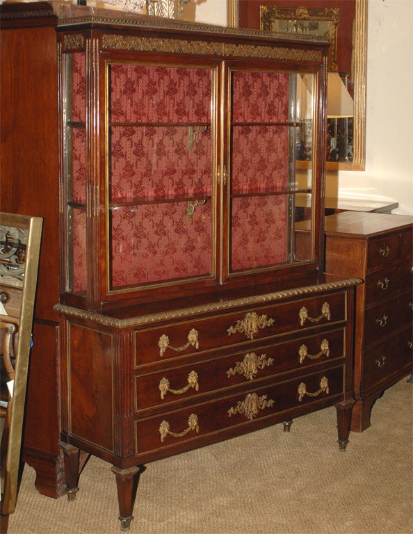 With ornate frieze above rectangular case; over paired glass doors revealing two interior shelves; mounted to base cabinet with three long drawers; embellished with ormolu hardware; the whole raised on tapered legs.