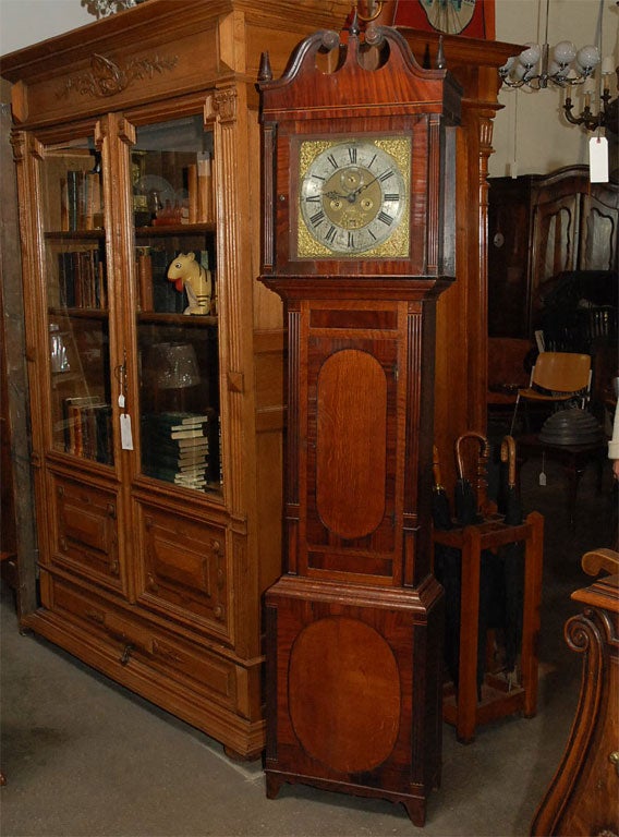 This eight day long case clock strikes the hours on a bell. It has: a square dial with silver chapter ring, brass mask corner spandrels, pierced hands, seconds hand, day indicator, pendulum and two iron weights. The door and base have central