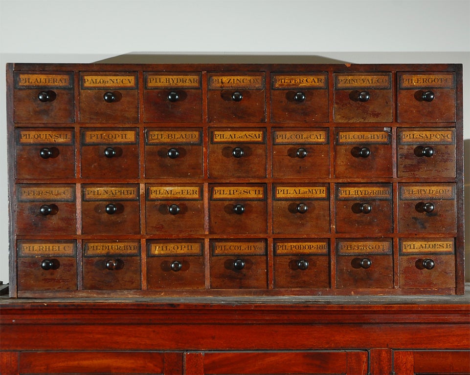 Early 19th century English apothecary wall chest with hand written labels, to hang or place on stand. The narrow rectangular body holds 28 square drawers with wooden knob handles.