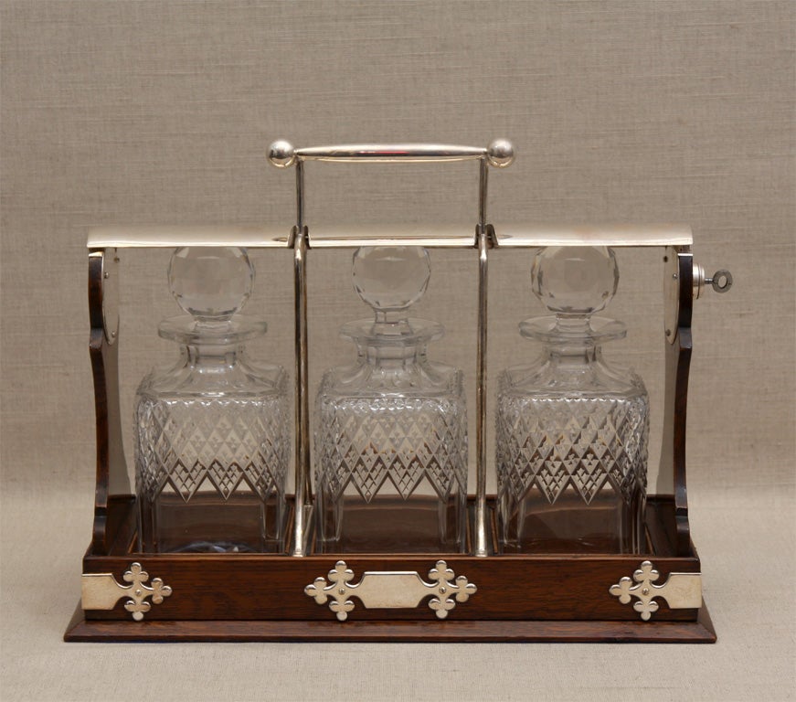 Oak and Crystal Tantalus Set with Three Cut-Crystal Decanters, Locking Silver Cross Bar, Key, and Handled Oak Stand Ornamented with Silver Monogram Plates.<br />
England, Late 19th Century.<br />
<br />
15.5 inches wide x 6 inches deep x 12