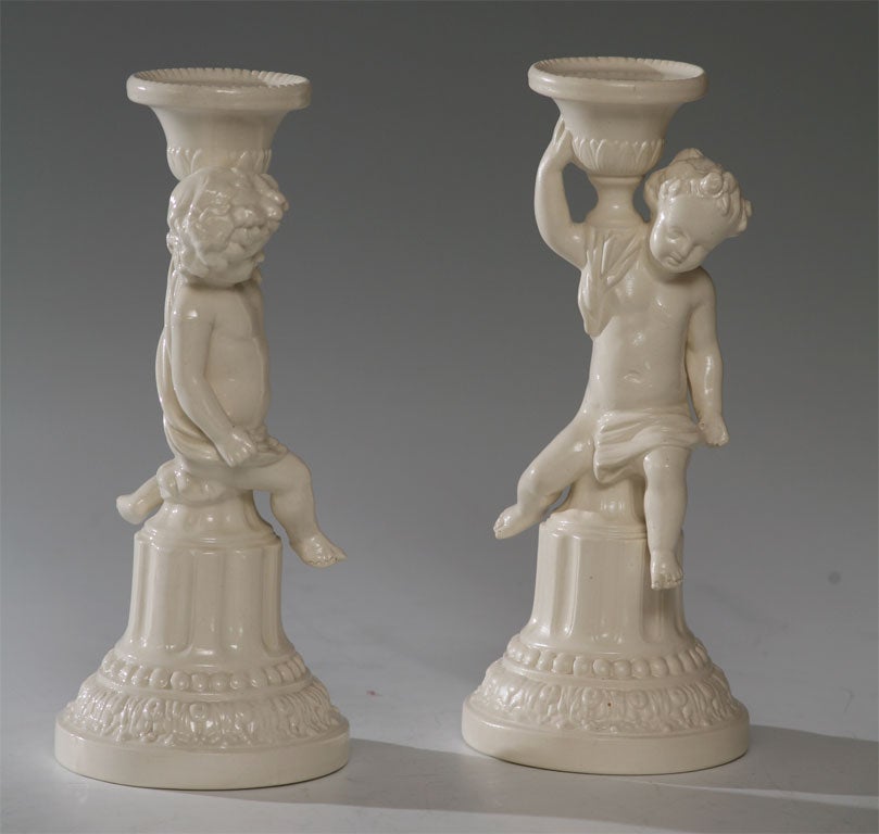 Matched pair of porcelain figural candlesticks with over-all white salt-glazed body. Classical Putti figures will embellish a country table setting or grace a mantle or sideboard.