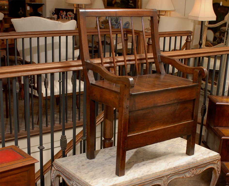 This unusual late 19th English chestnut chair features a curved comb back with spindles and a scrolling top rail, resting on four block feet with pegged construction. The arms, curved halfway, end in scrolls while a long carved apron joins both