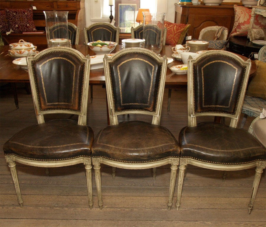 Six Louis XVI style dining chairs in faded black leather with gold tooling, nailhead trim and fluted legs. Crème paint with greige paint glaze.