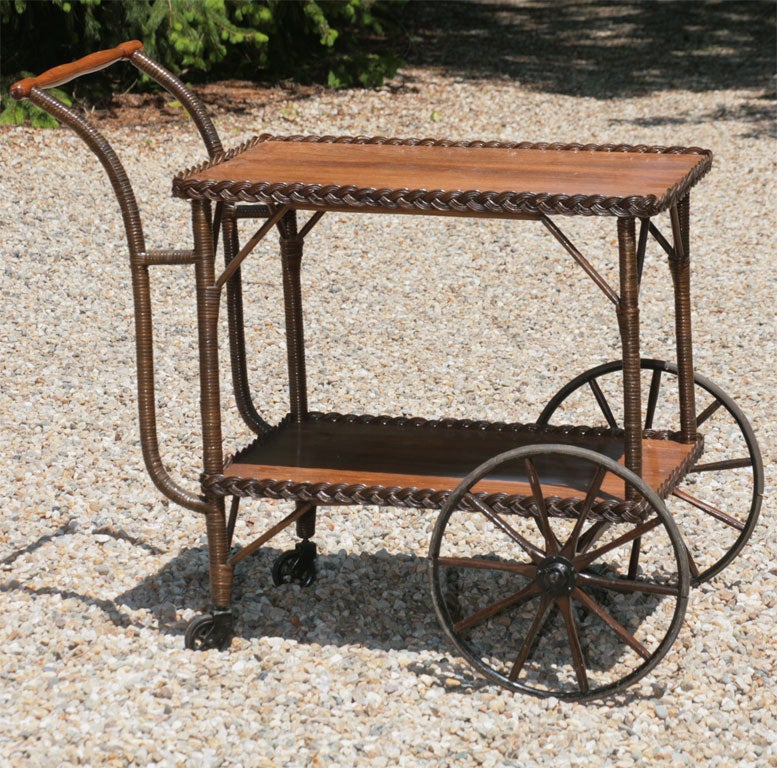 Classic wicker tea cart in natural stained finish having 2 solid wood surfaces finished in wicker braiding.  Wooden spoked front wheels, wood casters on back legs.  Gently curved cane-wrapped posts leading to turned wood handle.