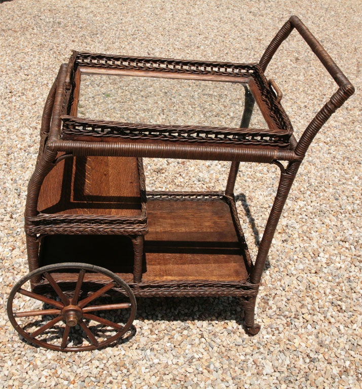 Victorian wicker tea cart in natural stained finish.  Removable wicker framed glass serving tray.  Full wooden bottom shelf and interior half shelf with woven galleries.  Wood spoked front wheels and wood casters on back legs.