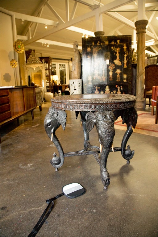 Intricately carved table. Top with foliate design surrounding four deities. Four carved elephant heads with ivory tusks holding jars of oil. Serpents on stretchers heading toward bowl of water.