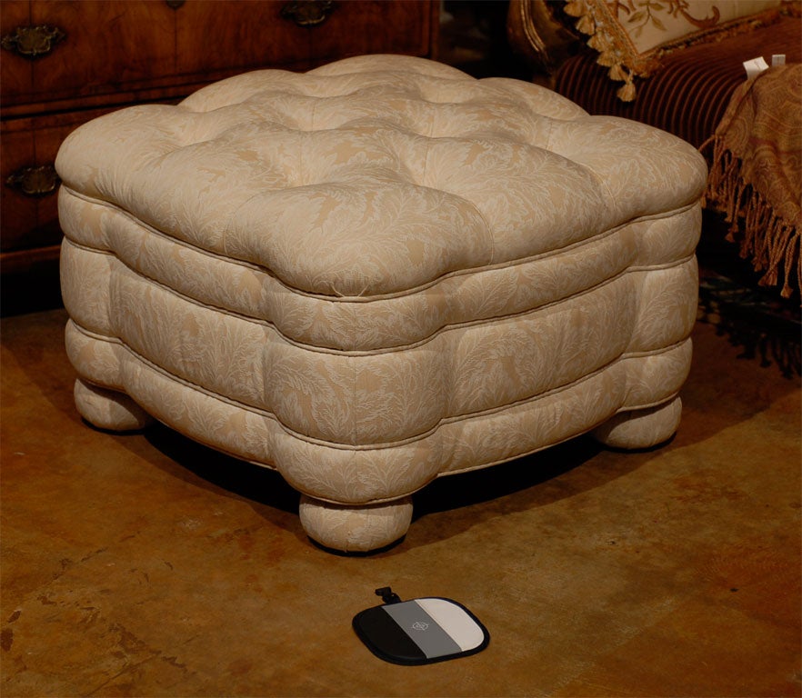 Lee Jofa ottoman upholstered in a cream and white damask fabric.  Ottoman has a tufted top, scalloped shape, and gimped decorative banding. The ottoman rests on upholstered bun feet.
