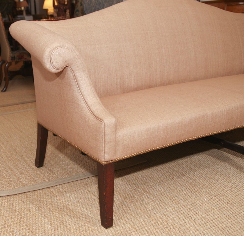The camel-back sofa with scrolled arms upholstered in close-nailed linen and on a mahogany frame joined by a stretcher.