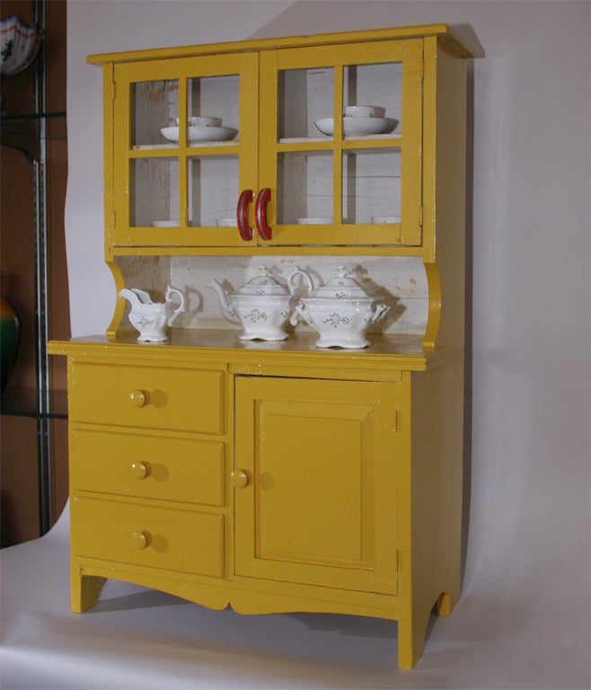 Vintage yellow painted child's cupboard with enclosed shelves over drawers and a cubby. Displayed on working space and in shelves is an English Staffordshire 