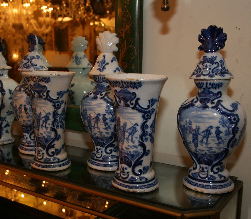 A five piece Dutch Delft garniture comprised of three covered vase and two open vase with flaring rims. The hand painted decoration in blue on a white ground depicting figures ice fishing. The bases having Greek frets, border detail, and urn with