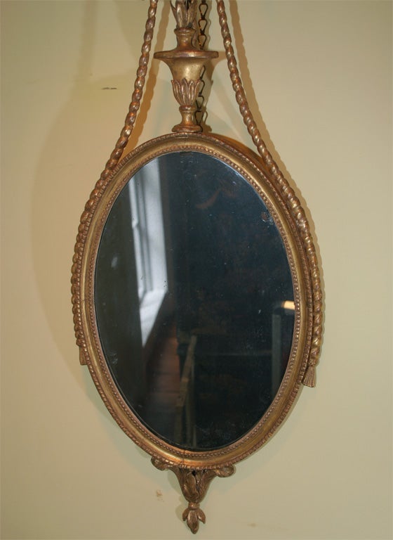 Very fine carved giltwood Adam mirror with carved rope and ribbon ornament centered by a patera above a classical urn with flowers. The oval mirror surround with beaded border, the bottom with incurvate leaf pendant ornament.
English, circa 1775.


