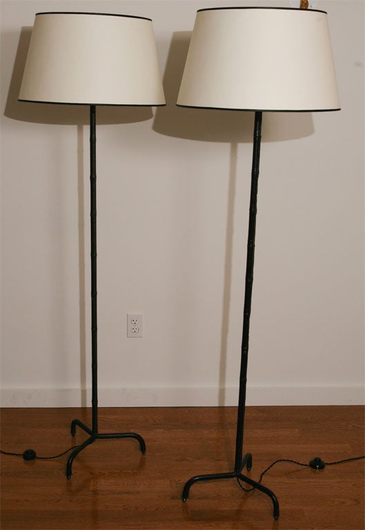 Mid-century Jacques Adnet floorlamps in original black leather on tripod base. Contrasting cream-stitched bamboo detail in leather. Floorlamps include white paper shades with black top and bottom trim. US rewired with black cotton cord.