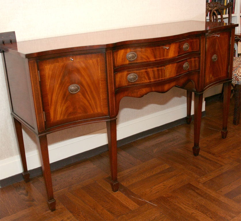 Georgian-Style Flame Mahogany Bowfront Sideboard with Boxwood Inlay Containing Two Fitted Cupboards with Locks and Two Center Drawers with Locks, Top Drawer Felted and Fitted for Flatware. Made in England in the Early - Mid 20th Century.<br />
<br