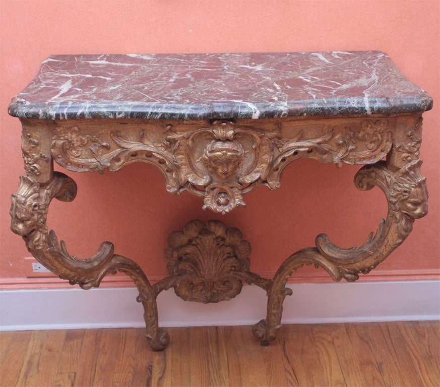 A carved and gilded wood console with a pierced waved foliate-carved frieze centering a mask of Venus over a pair of scrolled supports headed by lion’s masks and joined by a stretcher centering a ruffled shell, the sides carved with wings; all