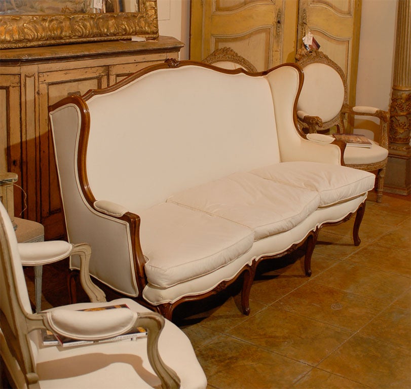 A French Louis XV style three-seat wooden winged sofa from the 19th century with carved crest, scrolled arms, cabriole legs and upholstery. This French wooden canapé à oreilles features a delicately curved upper rail accented in its center with a