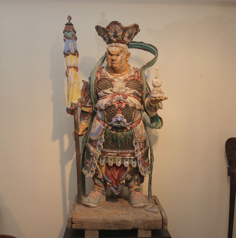 Large crowned Warrior King with a wrathful expression, bearing a stupa in his left hand, holding an umbrella with his right. Adorned with armor and flowing scarf.