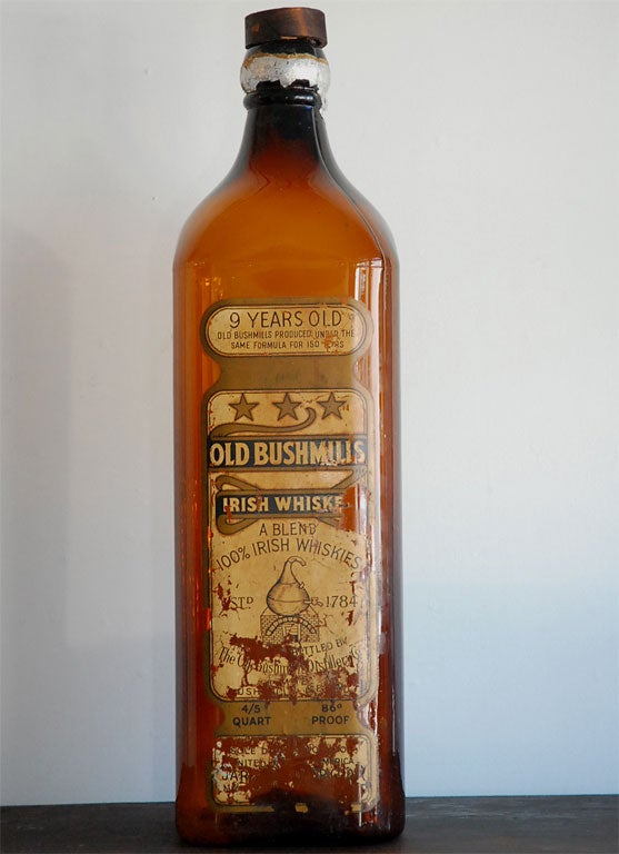 19TH C. OLD BUSHMILLS IRISH WHISKEY LARGE GLASS BOTTLE USED AS A TRADE SIGN AT A SALOON.  ORIGINAL LABEL IN TACT.