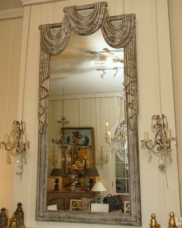 A dramatic and grand mercury glass mirror, the frame being of carved wood, the top of which is carved in the form of swags cascading down the sides. It is painted grey with some gesso showing through the paint. The mirror is in three pieces.