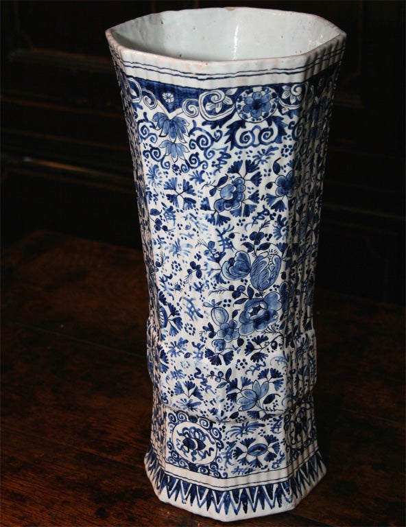 A tall, octagonal 17th century Dutch lily vase, having intricate hand-painted decoration depicting flowers, leaves and vines in varying hues of blue on a greyish white background, with striated and outstepped sides and a flaring top.