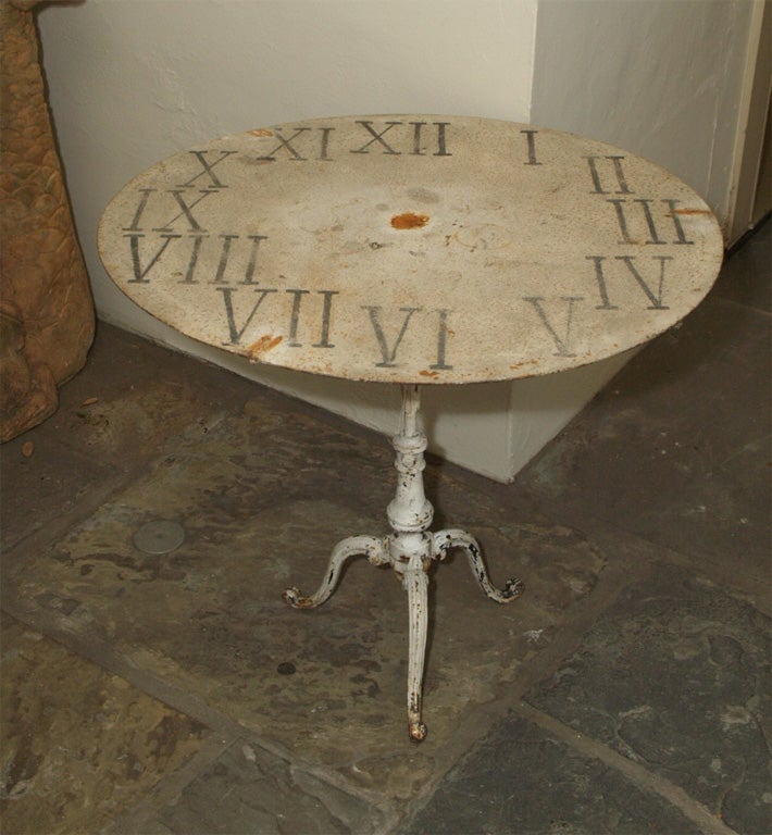 A GARDEN TABLE MADE FROM A CLOCK FACE. THE BASE IS OF BAULSTER FORM WITH 3 GRACEFULLY CURVED LEGS.  THREE CURVED ARMS SUPPORT THE TOP.