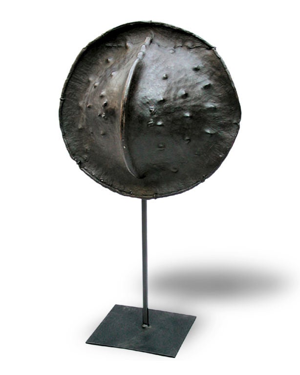 A LARGE AND UNUSUAL AFRICAN HIPPOPOTAMUS-SKIN CIRCULAR SHIELD. The well-patinated convex circular shield with ribbed middle tapering to a flared edge; restorations and replacements to include a later iron stand; wear and age cracks.<br />
A related