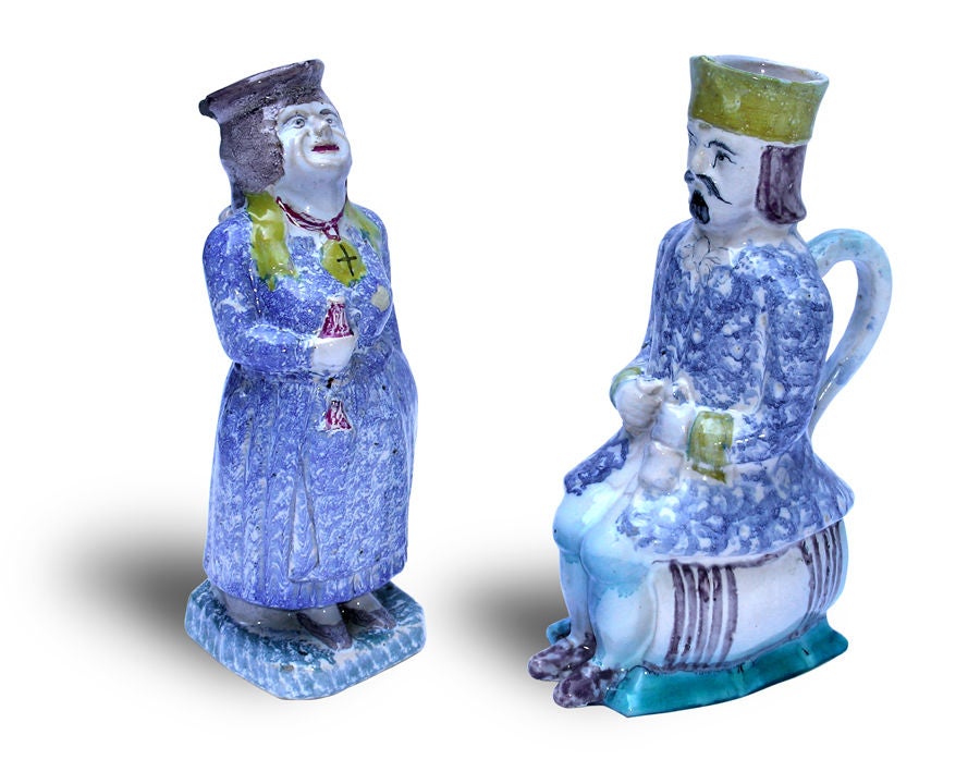Each capacious tankard of figural form with open cap; one, a TWO MIRTHFUL FRENCH POLYCHROMED FAIENCE FIGURAL TANKARDS WITH STRAP HANDLES. standing, laughing woman adorned with a cross-inscribed oval pendant and abbreviated shawl, and holding a small