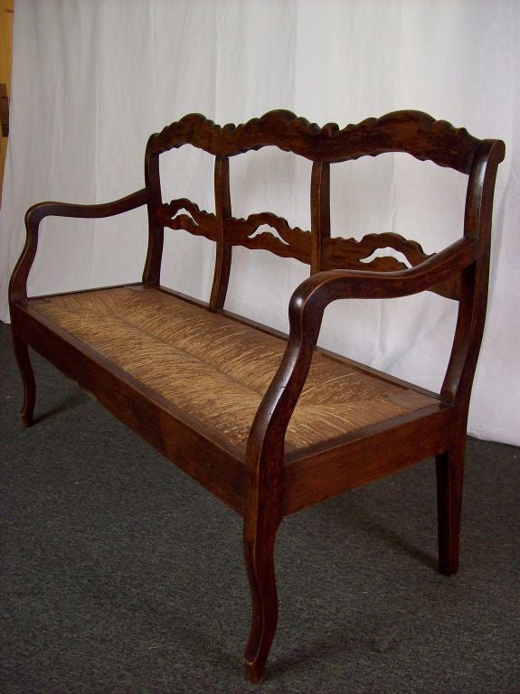 An antique bench from France with a rush seat. The gracefully curved arms and legs as well as the cutout shaped rails give this bench great charm. Made of fruitwood with a lovely color and patina. Old rush is in good condition. Very sturdy.