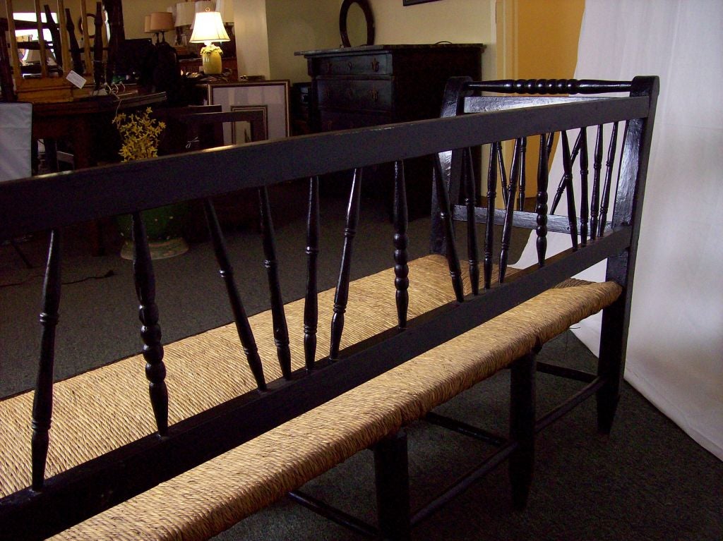 A charming antique bench from France with a very interesting profile,lovely curved arms, with a rush seat. In old black paint, with several details of note: turned legs and spindles, bobbin stretchers, and curved bobbin arm rails at top.