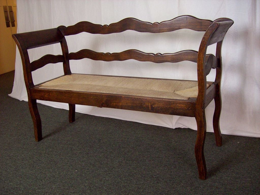An antique bench from France. Made of fruit wood, this bench has gracefully shaped rails with curved arms and legs. The old rush is in very good condition. Quite sturdy.