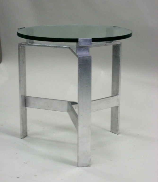 A pair of end or side tables in silver leafed solid metal in the style of Jacques Quinet. Each table is supported by a tripod base with each side cross-bracing the other. A round of glass is cantilevered above the frame to form the
