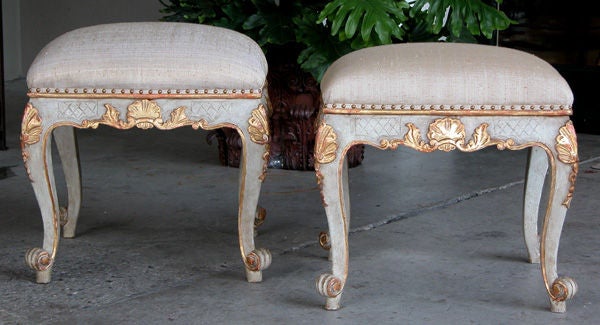 An elegant pair of Italian rococo style pale blue painted and parcel-gilt stools; each with serpentine-shaped upholstered seat above a scalloped apron with shell and foliate carving; raised on cabriole supports ending in a scrolled foot