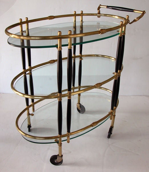 A chic French 1940's oval brass drinks cart with ebonized highlights and 3 glass shelves; consisting of 3 oval glass shelves within a brass frame raised on ebonized columnar supports; all over 4 bakelite casters