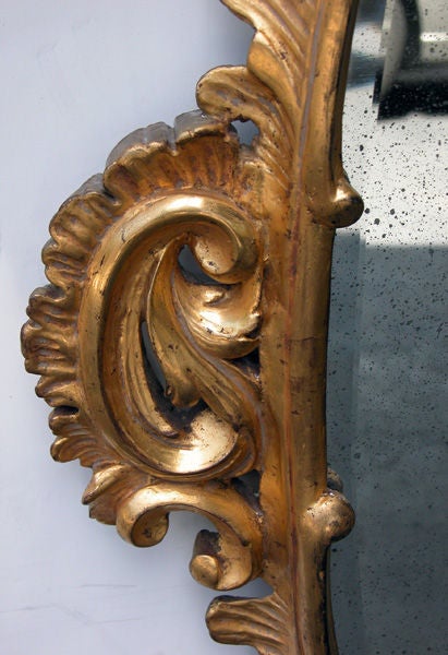 Miroir ovale en bois doré rococo de style Louis XV, élégamment sculpté, surmonté d'une crête rocaille ajourée au-dessus d'un cadre ovale orné d'une sculpture de feuillage avec des rinceaux en C exagérés sur les côtés et un pendentif de feuillage à