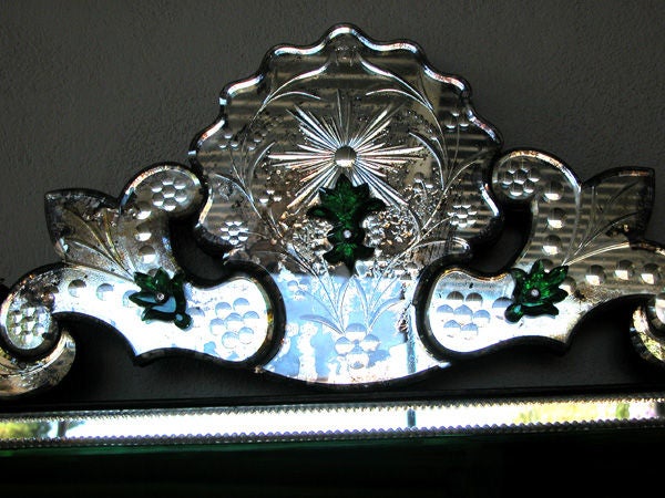 A shimmering and unusual Venetian mirror with emeral green glass frame; the arching etched mirrored crest centering a sylized flower head flanked by lively scrollwork; above a rectangular mirror plate enclosed by a clear and emerald green glass